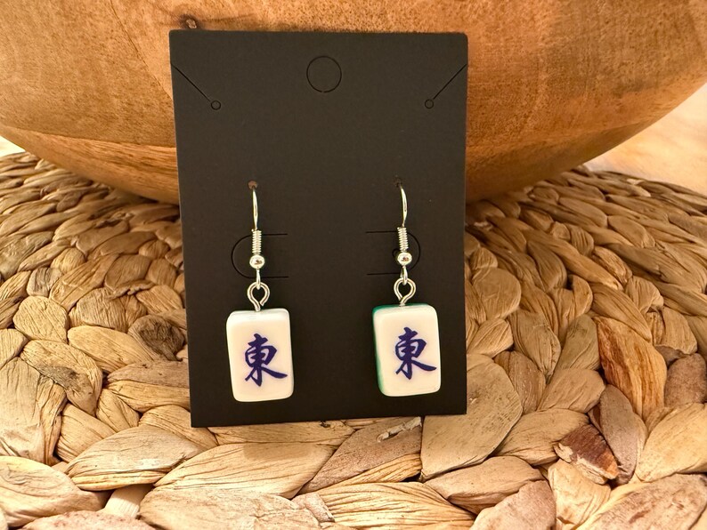 May include: A pair of dangle earrings featuring white mahjong tiles with the blue Chinese character for East, suspended from silver-toned metal hooks. The earrings are displayed on a rectangular black card with a circular hole at the top.