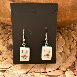 May include: A pair of dangle earrings featuring white rectangular mahjong tiles with the number 1 and a red floral design. The tiles are suspended from silver-toned metal fishhook wires and are displayed on a black rectangular card.