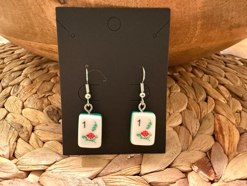 May include: A pair of dangle earrings featuring white rectangular mahjong tiles with the number 1 and a red floral design. The tiles are suspended from silver-toned metal fishhook wires and are displayed on a black rectangular card.