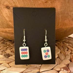 May include: A pair of drop earrings featuring white rectangular mahjong tiles with colorful circular patterns. The left earring displays a vertical column of blue, red, and green circles, while the right earring shows five circles in a quincunx arrangement.