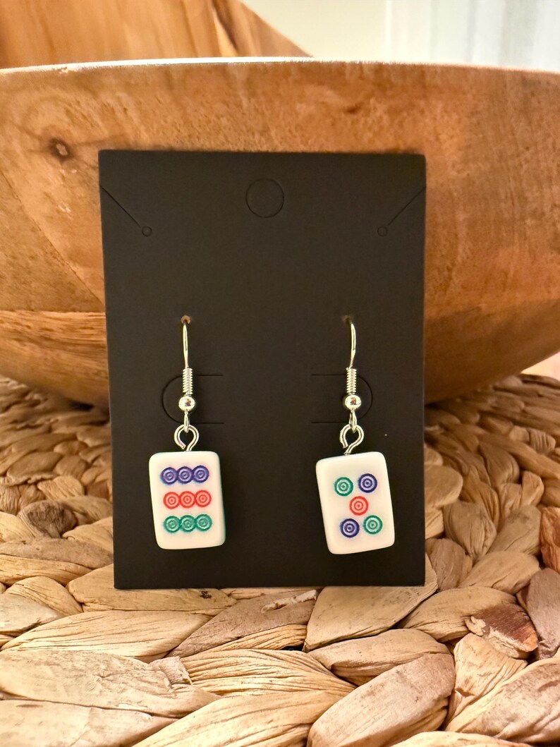 May include: A pair of drop earrings featuring white rectangular mahjong tiles with colorful circular patterns. The left earring displays a vertical column of blue, red, and green circles, while the right earring shows five circles in a quincunx arrangement.