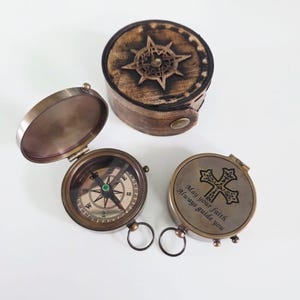 May include: Two brass pocket compasses and a round leather case. One compass is open, showing a directional dial with cardinal points. The other compass is closed, displaying an engraved cross and the text, May your faith Always guide you.