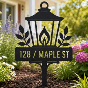 Pode incluir: Placa de endereço de metal preto em forma de lanterna, com o endereço 128 / MAPLE ST. A placa tem um design de chama e detalhes de folhas. Está colocada em um jardim.