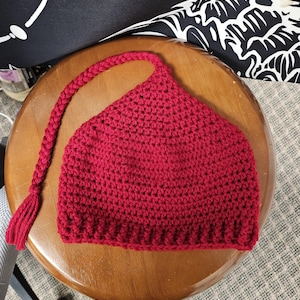 May include: A handmade, red crochet hat with a long, braided strap and tassel. The hat is sitting on a wooden stool. The hat is a solid, vibrant red color.