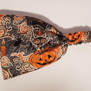 May include: A Halloween-themed headband with a gray background and orange, black, and brown patterns. The design includes pumpkins, spiders, spiderwebs, and a candle. The headband is made of fabric and has an elastic band.