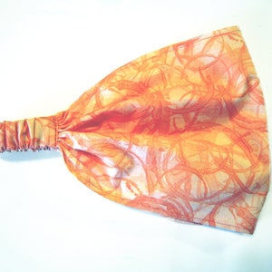 May include: Orange and yellow patterned headband made of fabric. The headband is elastic and has a gathered edge.