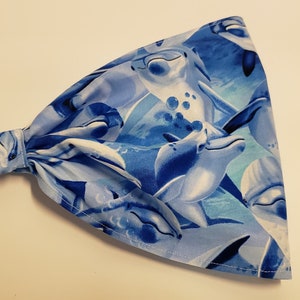 May include: A wide headband featuring a repeating pattern of dolphins in shades of blue and white. The fabric is gathered at the back with elastic for a comfortable fit. The headband is designed to keep hair out of the face.