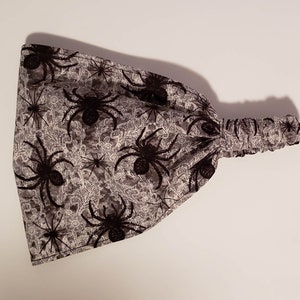 May include: A gray and white headband with a spider pattern. The headband features a repeating pattern of black spiders on a gray background with a white floral design. The elasticated section is gray.