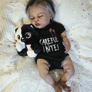 Puede incluir: Una mu&ntilde;eca beb&eacute; realista con cabello oscuro y un body negro que dice "CAREFUL | BITE!". La mu&ntilde;eca sostiene un panda de peluche blanco y negro. La mu&ntilde;eca est&aacute; acostada sobre una manta blanca acolchada.