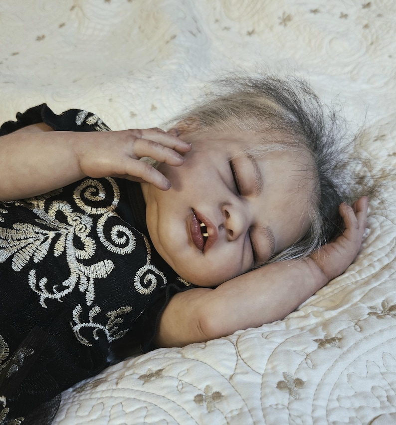Puede incluir: Una mu&ntilde;eca beb&eacute; realista con los ojos cerrados, acostada sobre una manta crema estampada. La mu&ntilde;eca lleva un vestido negro con bordados florales blancos. El pelo de la mu&ntilde;eca es gris y negro, y tiene dos dientes visibles.