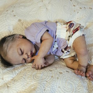 Puede incluir: Una mu&ntilde;eca beb&eacute; realista est&aacute; acostada sobre una manta blanca estampada. La mu&ntilde;eca lleva una camiseta de manga corta de color lila claro y una braguita con estampado floral con un lazo grande. La mu&ntilde;eca tiene pelo oscuro con una raya blanca y est&aacute; durmiendo.