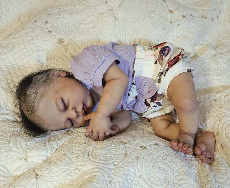 Puede incluir: Una mu&ntilde;eca beb&eacute; realista est&aacute; acostada sobre una manta blanca estampada. La mu&ntilde;eca lleva una camiseta de manga corta de color lila claro y una braguita con estampado floral con un lazo grande. La mu&ntilde;eca tiene pelo oscuro con una raya blanca y est&aacute; durmiendo.