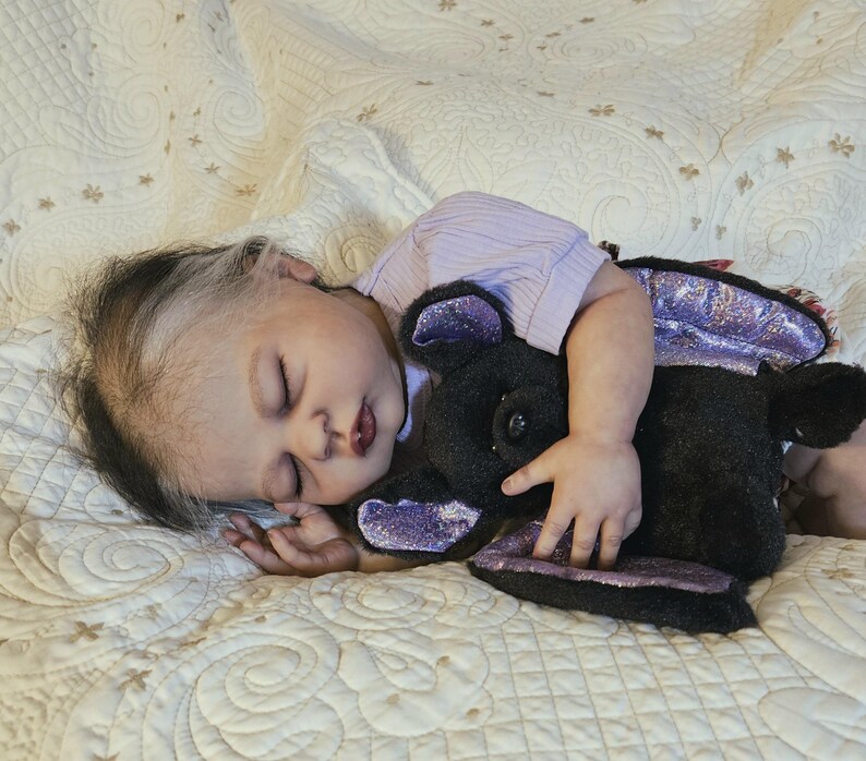 Puede incluir: Una mu&ntilde;eca beb&eacute; durmiente con cabello oscuro y una camisa lila claro, abrazando un peluche negro con detalles morados. La mu&ntilde;eca est&aacute; acostada sobre una manta blanca acolchada con un patr&oacute;n floral.