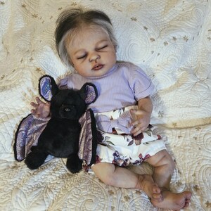 Puede incluir: Una mu&ntilde;eca beb&eacute; realista con los ojos cerrados, vestida con una camiseta lavanda de manga corta y una falda con estampado floral. Un murci&eacute;lago de peluche negro con alas moradas descansa junto a la mu&ntilde;eca sobre una manta acolchada.