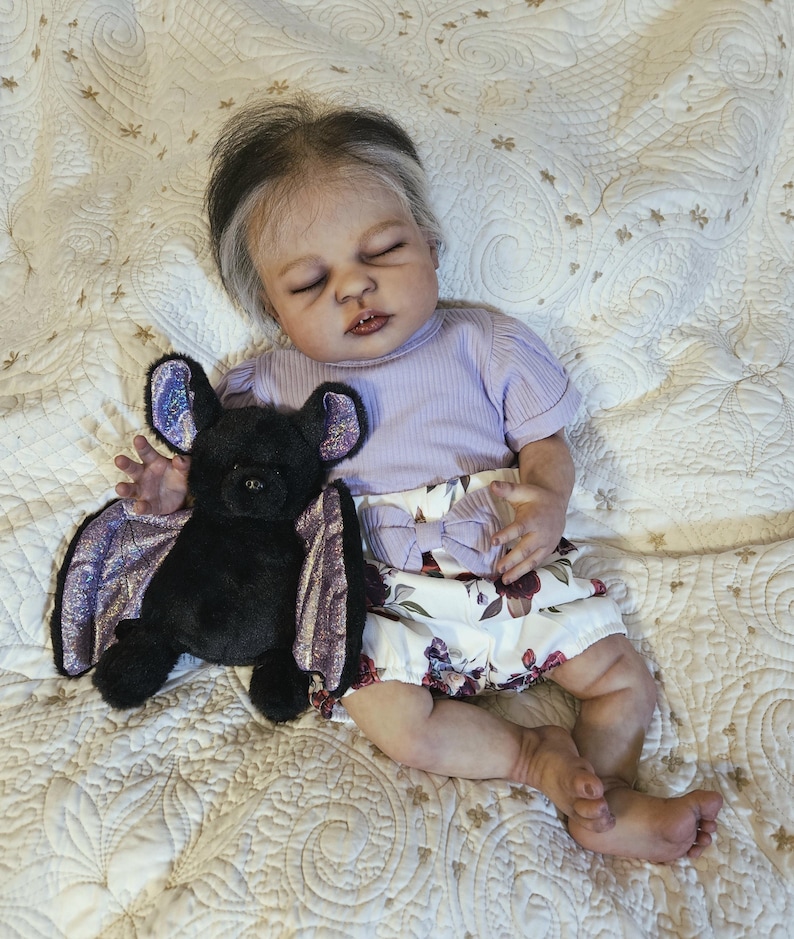 Puede incluir: Una mu&ntilde;eca beb&eacute; realista con los ojos cerrados, vestida con una camiseta lavanda de manga corta y una falda con estampado floral. Un murci&eacute;lago de peluche negro con alas moradas descansa junto a la mu&ntilde;eca sobre una manta acolchada.