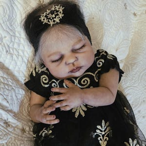 Puede incluir: Una mu&ntilde;eca beb&eacute; realista con los ojos cerrados, vestida con un vestido negro con bordados dorados. La mu&ntilde;eca tiene cabello oscuro con un adorno para la cabeza y est&aacute; acostada sobre una superficie acolchada blanca.