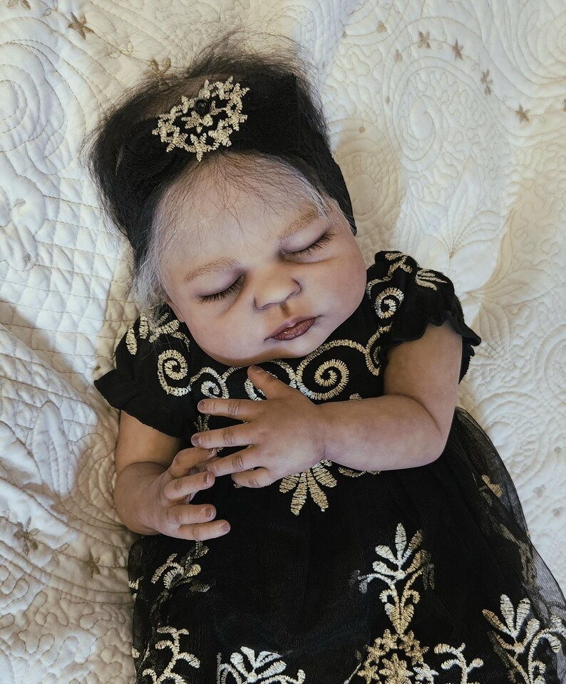 Puede incluir: Una mu&ntilde;eca beb&eacute; realista con los ojos cerrados, vestida con un vestido negro con bordados dorados. La mu&ntilde;eca tiene cabello oscuro con un adorno para la cabeza y est&aacute; acostada sobre una superficie acolchada blanca.