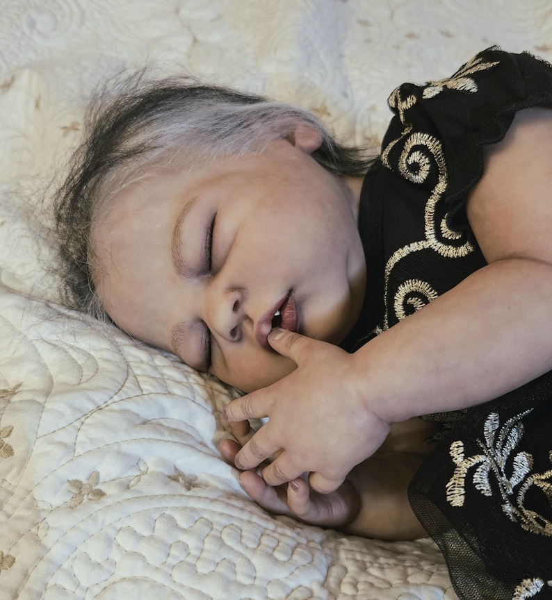 Puede incluir: Una mu&ntilde;eca beb&eacute; realista con los ojos cerrados, descansando sobre una superficie acolchada de color crema. La mu&ntilde;eca tiene la piel clara, cabello oscuro con un toque de blanco y lleva un vestido negro con bordados dorados. La mano de la mu&ntilde;eca est&aacute; cerca de su boca.