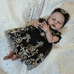 Puede incluir: Una mu&ntilde;eca beb&eacute; realista con un vestido negro con bordados florales dorados. La mu&ntilde;eca tiene la piel clara, los ojos cerrados y una diadema negra con un dise&ntilde;o floral a juego. La mu&ntilde;eca est&aacute; acostada sobre una manta blanca acolchada.