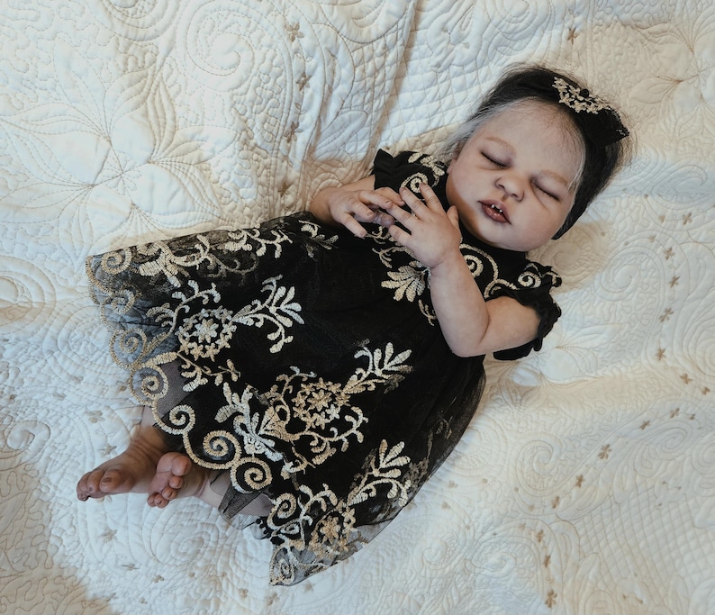 Puede incluir: Una mu&ntilde;eca beb&eacute; realista con un vestido negro con bordados florales dorados. La mu&ntilde;eca tiene la piel clara, los ojos cerrados y una diadema negra con un dise&ntilde;o floral a juego. La mu&ntilde;eca est&aacute; acostada sobre una manta blanca acolchada.