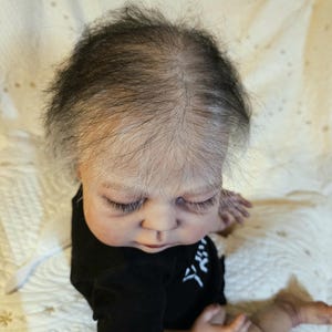 Puede incluir: Una mu&ntilde;eca reborn realista con tez clara, ojos cerrados y pesta&ntilde;as finas. La mu&ntilde;eca lleva un body negro con caracteres japoneses blancos. El pelo de la mu&ntilde;eca es una mezcla de negro y gris, sobre una superficie blanca.