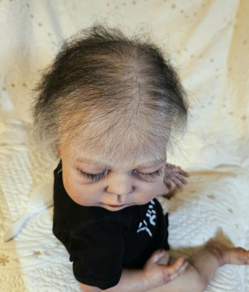 Puede incluir: Una mu&ntilde;eca reborn realista con tez clara, ojos cerrados y pesta&ntilde;as finas. La mu&ntilde;eca lleva un body negro con caracteres japoneses blancos. El pelo de la mu&ntilde;eca es una mezcla de negro y gris, sobre una superficie blanca.