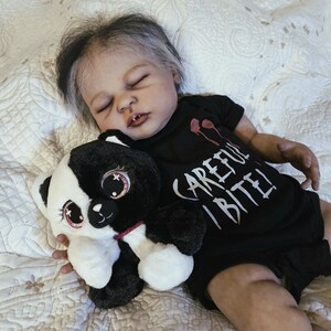 Puede incluir: Una mu&ntilde;eca beb&eacute; realista con cabello gris y negro, vestida con un body negro con el texto "CAREFUL BITE!". La mu&ntilde;eca sostiene un panda de peluche blanco y negro con detalles en rosa. La mu&ntilde;eca est&aacute; acostada sobre una superficie acolchada blanca.