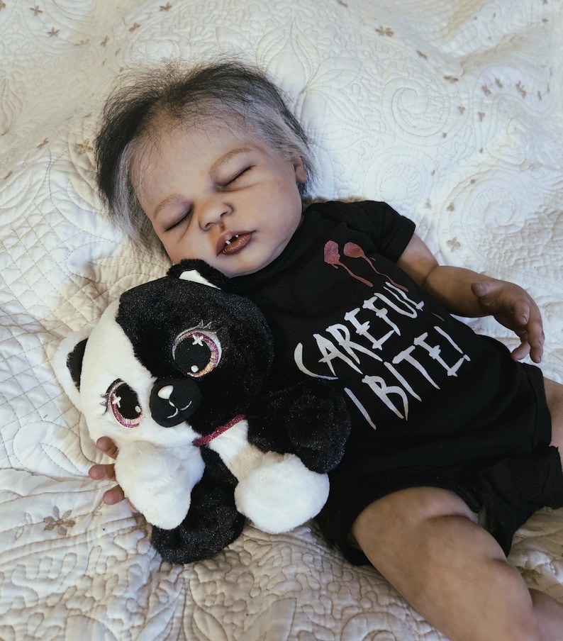Puede incluir: Una mu&ntilde;eca beb&eacute; realista con cabello gris y negro, vestida con un body negro con el texto "CAREFUL BITE!". La mu&ntilde;eca sostiene un panda de peluche blanco y negro con detalles en rosa. La mu&ntilde;eca est&aacute; acostada sobre una superficie acolchada blanca.