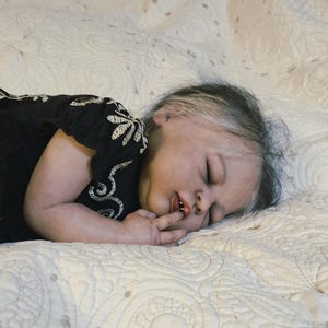 Puede incluir: Una mu&ntilde;eca beb&eacute; realista con los ojos cerrados, acostada sobre una manta acolchada de color crema. La mu&ntilde;eca lleva un vestido negro con bordados florales blancos. El pelo de la mu&ntilde;eca es oscuro con mechones blancos.