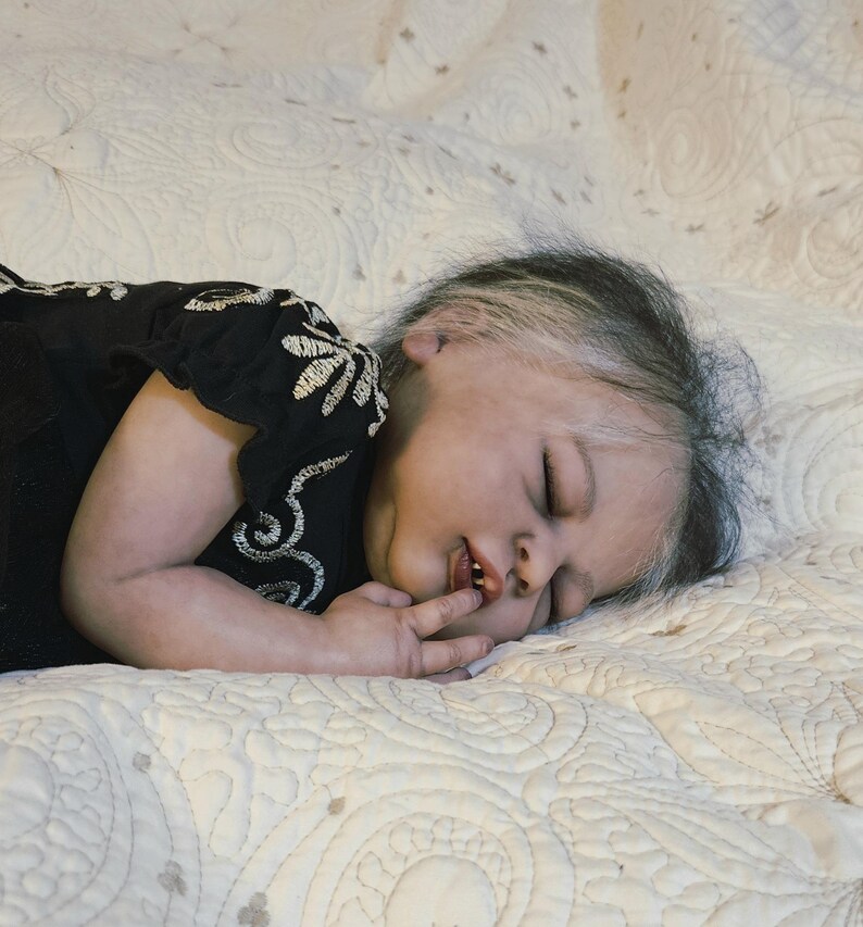 Puede incluir: Una mu&ntilde;eca beb&eacute; realista con los ojos cerrados, acostada sobre una manta acolchada de color crema. La mu&ntilde;eca lleva un vestido negro con bordados florales blancos. El pelo de la mu&ntilde;eca es oscuro con mechones blancos.