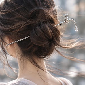May include: A silver-toned hair pin featuring a miniature faucet design at one end. The faucet is detailed with a cross-handle top and a small, clear, teardrop-shaped crystal charm hanging from the spout, creating a realistic water droplet effect.