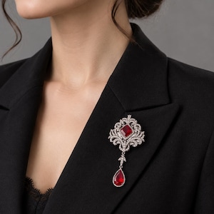 May include: A silver-toned brooch featuring a central square-cut red gemstone surrounded by clear crystals in an ornate filigree setting, with a dangling teardrop-shaped red gemstone pendant attached to the base of the main piece.