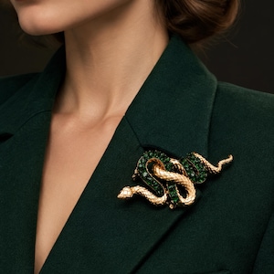 May include: A gold-toned snake brooch featuring a textured metallic body and segments inlaid with deep green crystals. The serpent is coiled in a winding shape, pinned to a dark green blazer, complemented by a matching oval green crystal earring.