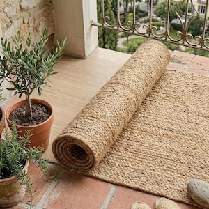 May include: A rolled natural fibre rug with a thick, braided texture sits on a tiled surface. The rug is light tan in colour and features a consistent woven pattern throughout. Beside the rug are two potted plants in terracotta-coloured pots.