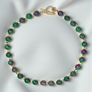Op de afbeelding: Een kralenketting met afwisselend ronde groene en magentakleurige zoisiet kralen en kleine, gefacetteerde goudkleurige metalen tussenkralen. De ketting wordt gesloten met een goudkleurige toggle-sluiting in de vorm van een hart.