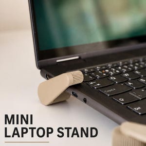 Può includere: Un supporto per portatile beige, compatto e a forma di cuneo, fissato al lato di un portatile nero. Il testo "MINI LAPTOP STAND" e "Compact support. Better posture. Anywhere." è stampato in nero sul lato sinistro dell'immagine.