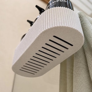 May include: An off-white, oval-shaped shower shelf with a ribbed side texture and a slatted base for drainage. The shelf is mounted to hold black pump bottles, one of which is clearly labeled with the word SHAMPOO and the word SILK underneath.