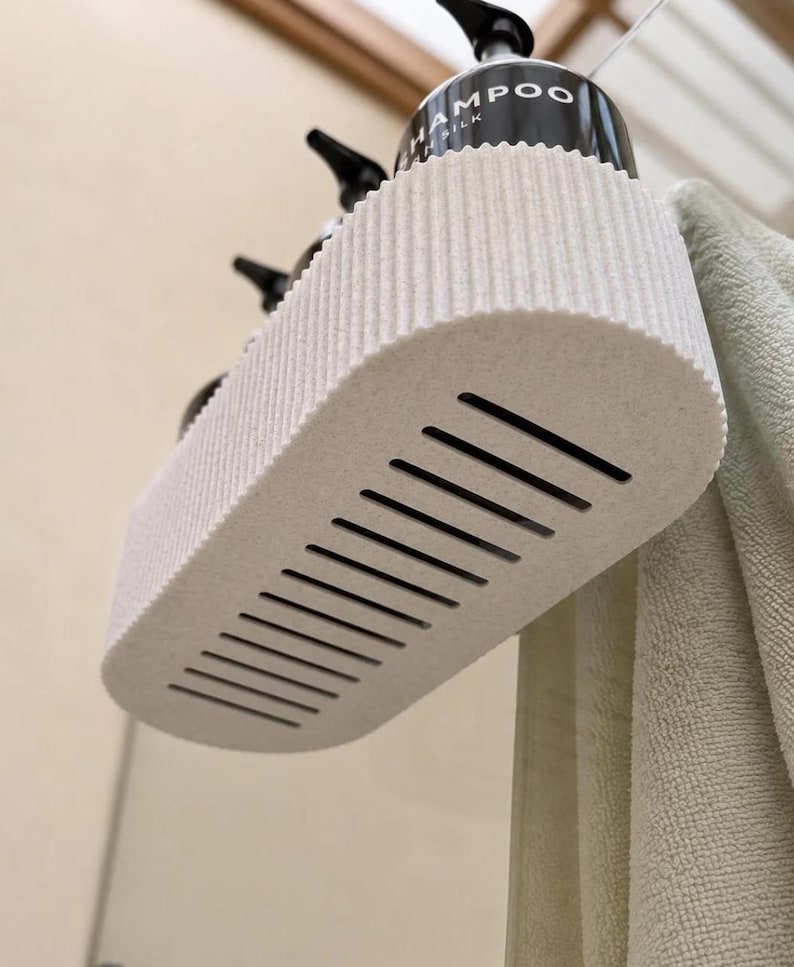 May include: An off-white, oval-shaped shower shelf with a ribbed side texture and a slatted base for drainage. The shelf is mounted to hold black pump bottles, one of which is clearly labeled with the word SHAMPOO and the word SILK underneath.