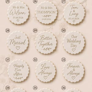 May include: A collection of twelve round, scalloped-edge white ceramic or clay discs arranged in a grid. Each disc features unique engraved text and symbols related to weddings, such as Mr & Mrs Wilson, Just Married, and Happily Ever After.