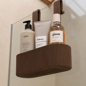 May include: A brown, oval-shaped shower caddy with a ribbed exterior and two hooks for hanging. It holds three personal care products: a Ouai scrub bottle, a tube of The Ordinary moisturizer, and a Gisou hair oil bottle with a light-colored cap.