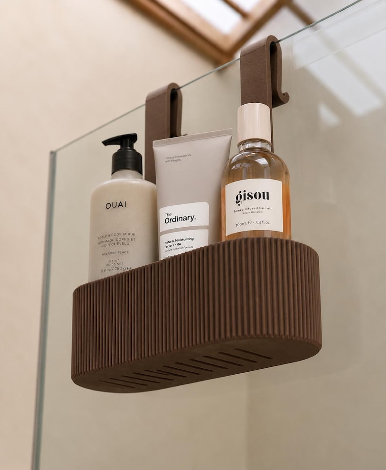 May include: A brown, oval-shaped shower caddy with a ribbed exterior and two hooks for hanging. It holds three personal care products: a Ouai scrub bottle, a tube of The Ordinary moisturizer, and a Gisou hair oil bottle with a light-colored cap.
