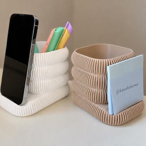May include: Two modern desk organizers. One is white and holds a smartphone and pens. The other is tan and holds sticky notes. Both have a ribbed, wavy design. The tan organizer has the text "@kenshohome" on the sticky notes.