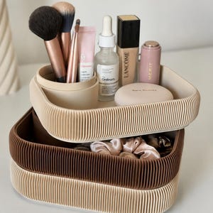 May include: Three beige and brown corrugated storage trays are stacked. The top tray contains makeup brushes, skincare products, and cosmetics. The bottom tray holds pink scrunchies. The trays are designed for cosmetic storage.