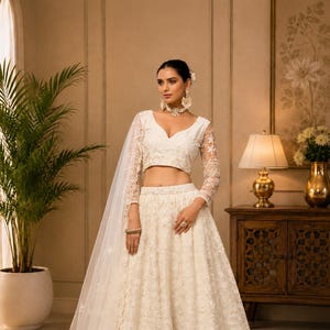 Könnte beinhalten: Eine elfenbeinfarbene, bestickte Lehenga mit einem passenden, langärmeligen, cropped Oberteil und einem transparenten Schleier. Der Rock ist weit und fließend, mit komplizierten Blumenmustern. Das Oberteil hat einen tiefen Ausschnitt und zarte Spitzärmel. Geeignet für besondere Anlässe.