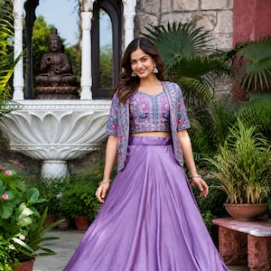 May include: A lavender floor-length skirt paired with a matching cropped top and a short-sleeved jacket. The top and jacket feature intricate embroidery with floral and bird motifs in shades of blue, pink, and green on a lavender fabric base.