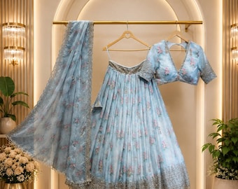 Könnte beinhalten: Ein hellblaues Lehenga-Set bestehend aus einem plissierten Rock und einer passenden Bluse mit tiefem V-Ausschnitt. Beide Teile sind mit einem zarten Blumenmuster und aufwendigen silbernen Stickereien am Saum verziert, ergänzt durch eine passende Dupatta.