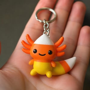 Op de afbeelding: Een sleutelhanger met een klein axolotl-figuurtje in de kleuren van maissnoepjes. De axolotl heeft een witte puntige kop, een oranje middengedeelte met drie kieuwen aan elke kant en een geel onderlichaam. Het heeft een gebogen staart en een metalen ring.
