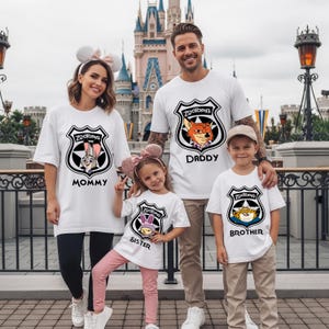 May include: A family of four wearing matching white t-shirts with Zootopia badge designs. The shirts feature the words "Mommy," "Daddy," "Sister," and "Brother." The background includes a castle and a decorative fence.