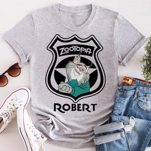 May include: A light gray t-shirt featuring a Zootopia badge design with the name "ROBERT" below. The badge includes the word "ZOOTOPIA" and a cartoon character. The shirt is paired with blue jeans and white sneakers.