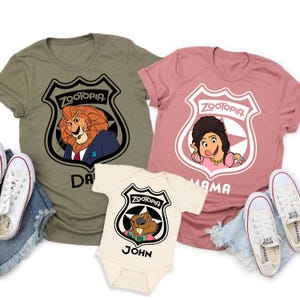 May include: Three Zootopia-themed t-shirts and a onesie. The shirts are olive green and dusty rose, and the onesie is cream-colored. Each features a Zootopia badge with a character image and a name. White sneakers are visible on either side of the shirts.